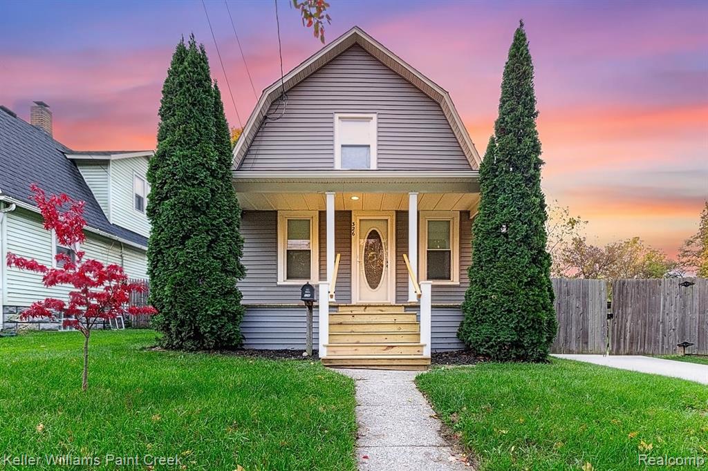 Gambrel-style exterior featuring grey siding, a covered front porch with a wood-finished staircase, and symmetrical evergreen landscaping