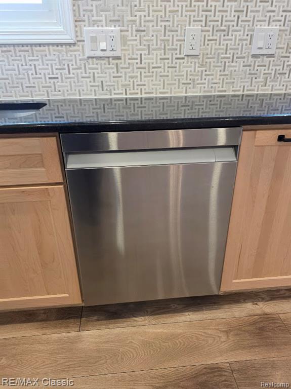 Kitchen view of stainless steel dishwasher, wood finished floors, tasteful backsplash, dark stone countertops, and light brown cabinetry