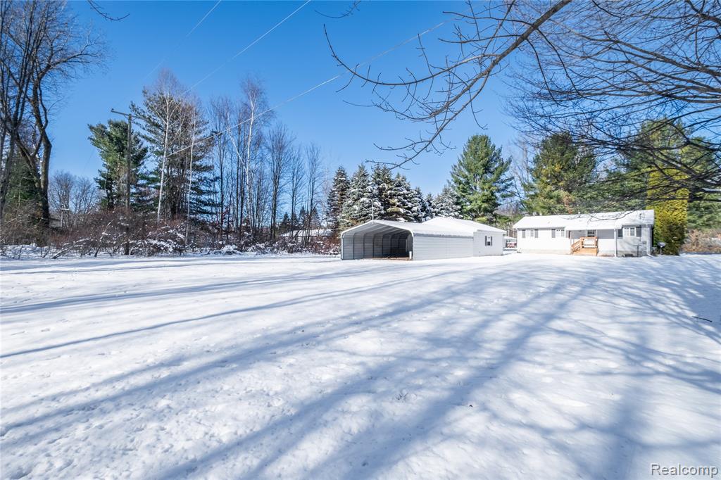 Yard layered in snow featuring a carport and view of wooded area