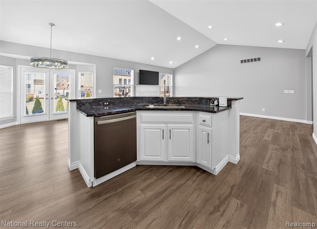 Kitchen with white cabinets, stainless steel dishwasher, dark stone countertops, dark wood-style flooring, and an island with sink
