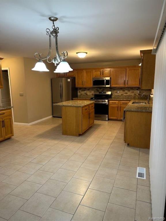 Kitchen featuring brown cabinets, appliances with stainless steel finishes, decorative light fixtures, dark stone countertops, and a center island