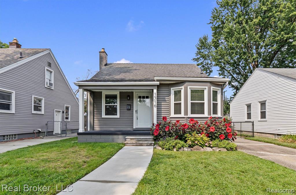 Gray siding exterior with white trim, covered front entry, bay window with louvered blinds, mature landscaping with flowering shrubs, and a concrete walkway