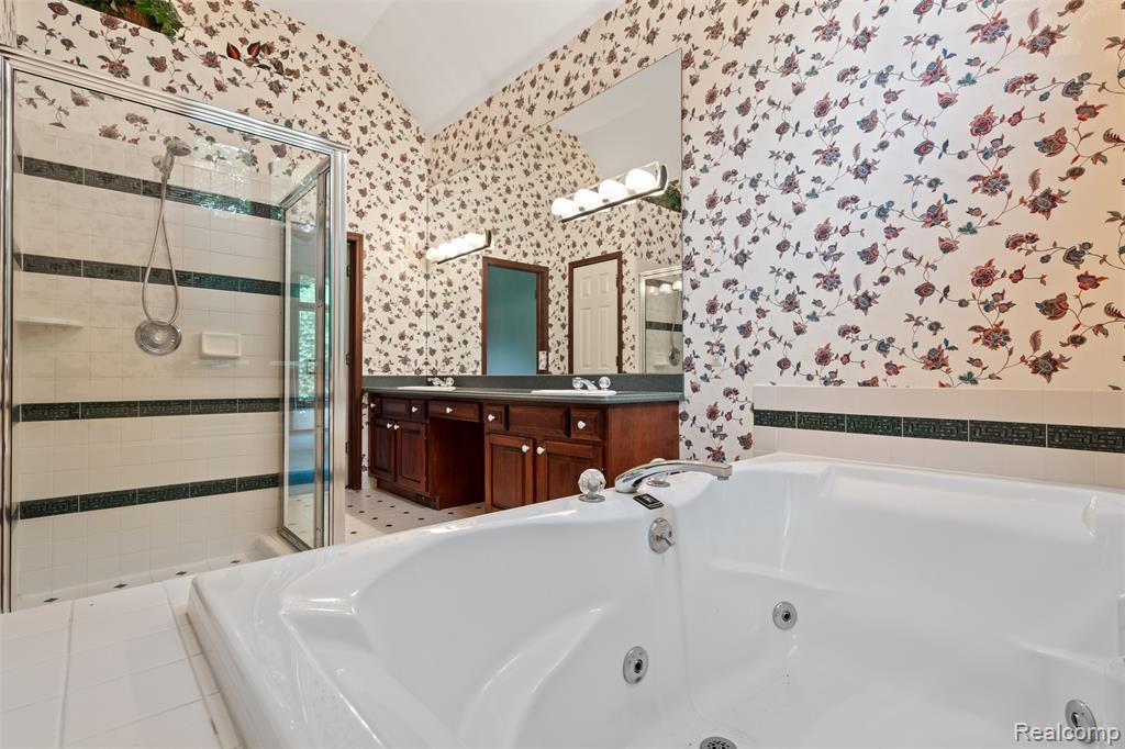 Full bath with a tub with jets, double vanity, wallpapered walls, lofted ceiling, and a stall shower