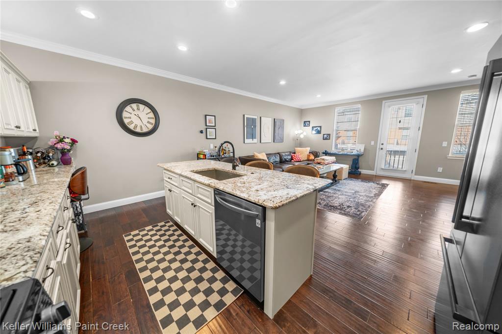 Kitchen featuring dark wood-type flooring, light stone countertops, dishwasher, freestanding refrigerator, and a center island with sink