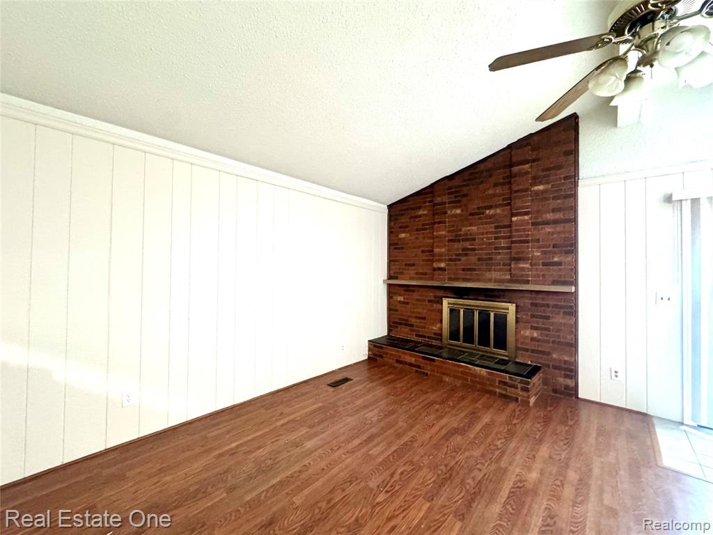 Unfurnished living room with a brick fireplace, wood finished floors, ceiling fan, lofted ceiling, and a textured ceiling