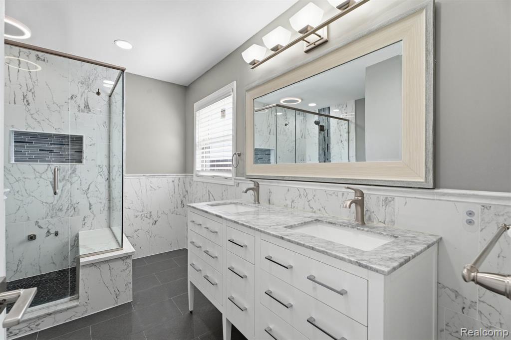 Bathroom with tile walls, double vanity, wainscoting, a marble finish shower, and recessed lighting