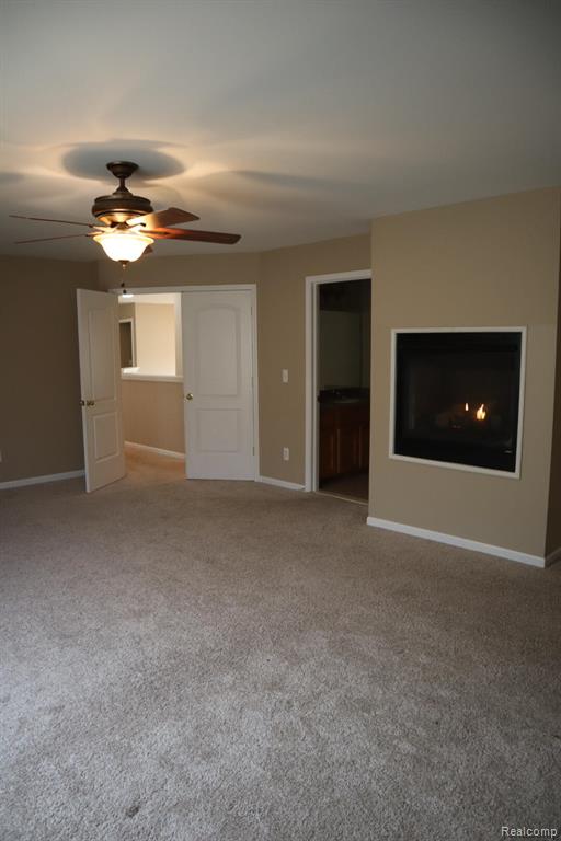 Unfurnished bedroom with ensuite bath, light colored carpet, a ceiling fan, and a glass covered fireplace