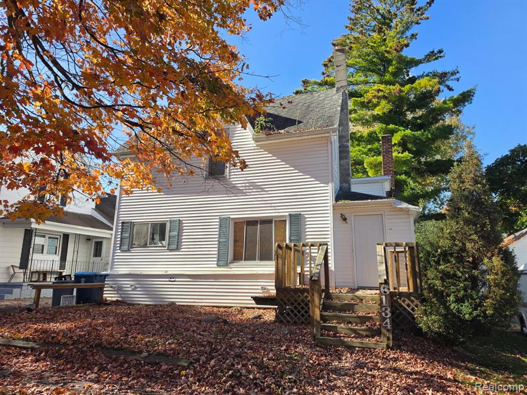 Two-story exterior featuring light-colored siding, dark shutters, a prominent brick chimney, and a wooden deck