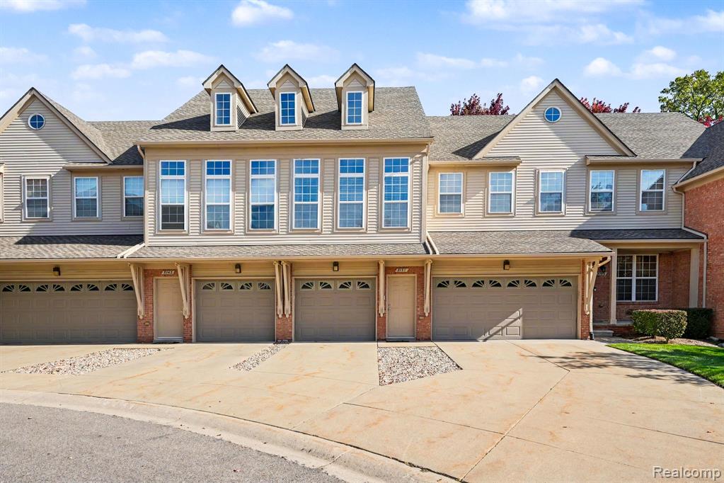 Multi-unit exterior featuring beige siding, tan garage doors with decorative arched window inserts, three roof dormers, and stone-accented concrete driveways