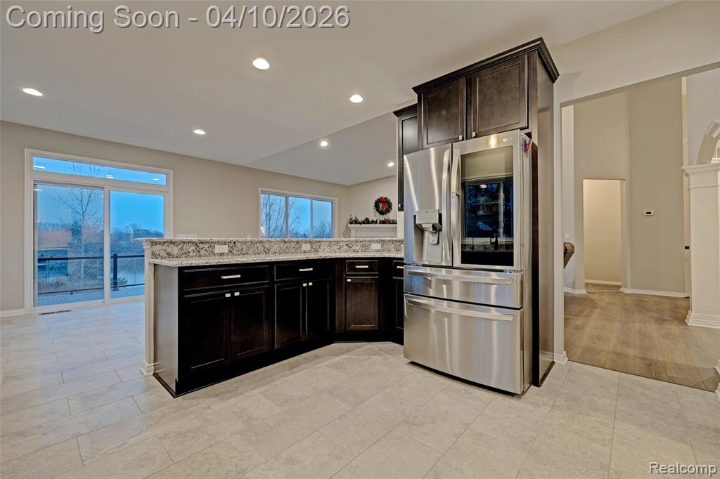 Kitchen with stainless steel fridge, a peninsula, dark wood finish cabinets, light stone counters, and recessed lighting