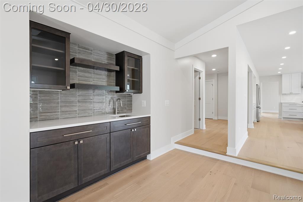 Great room wet bar featuring glass fronted cabinets, light wood-type flooring, decorative backsplash, dark wood finish cabinetry, and open shelves