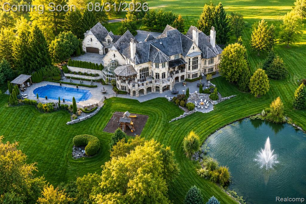 Stately stone exterior with multiple gables and dormers, expansive manicured lawn, inground swimming pool with surrounding patio, private pond with fountain, and mature landscaping