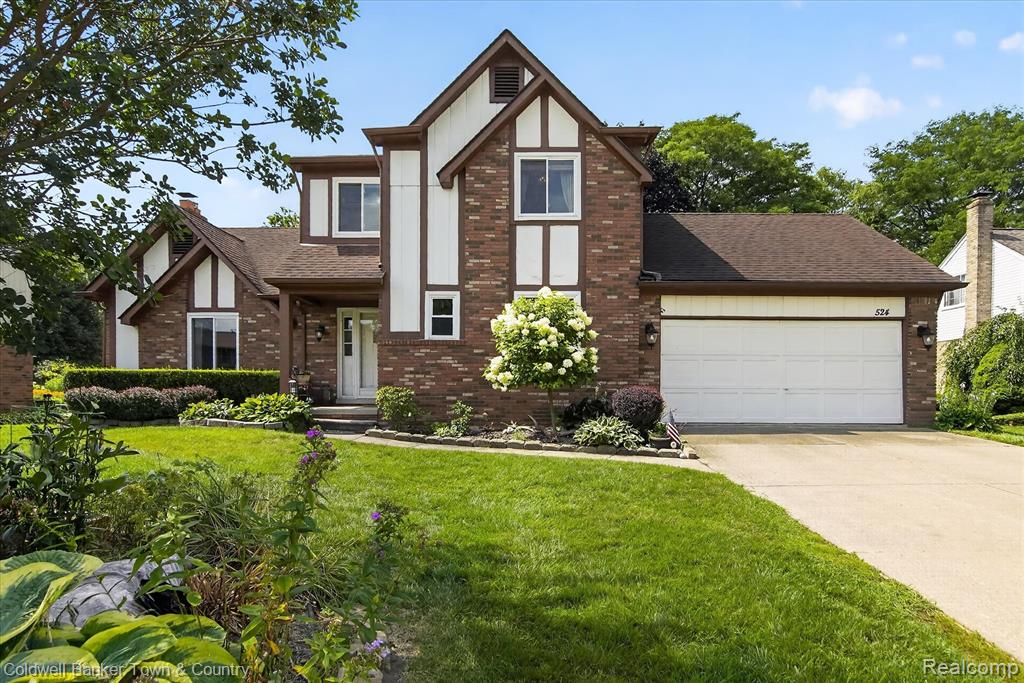 Brick exterior with white accents, two-car garage, mature landscaping, paved driveway, and covered front entry