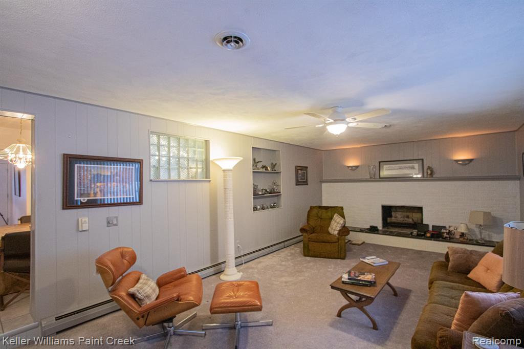 Living area with carpet flooring, a baseboard radiator, ceiling fan, wood walls, and a brick fireplace
