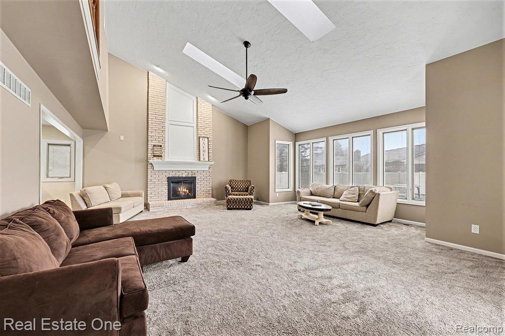 Living room featuring a brick fireplace, carpet flooring, high vaulted ceiling, a textured ceiling, and a skylight